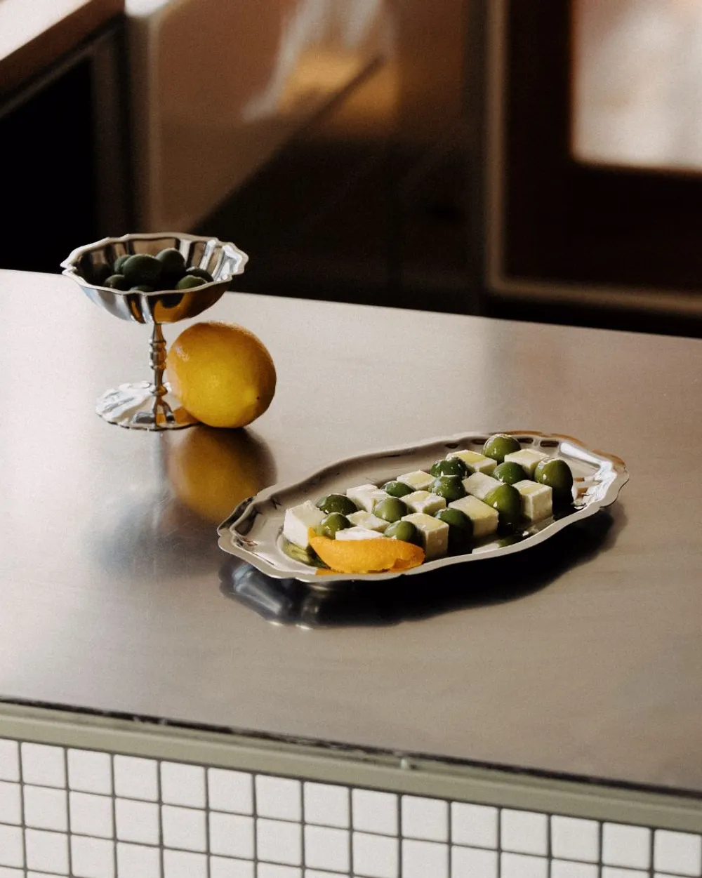 Riva stainless steel coupe of olives and plate of cheese and olives on a steel kitchen bench