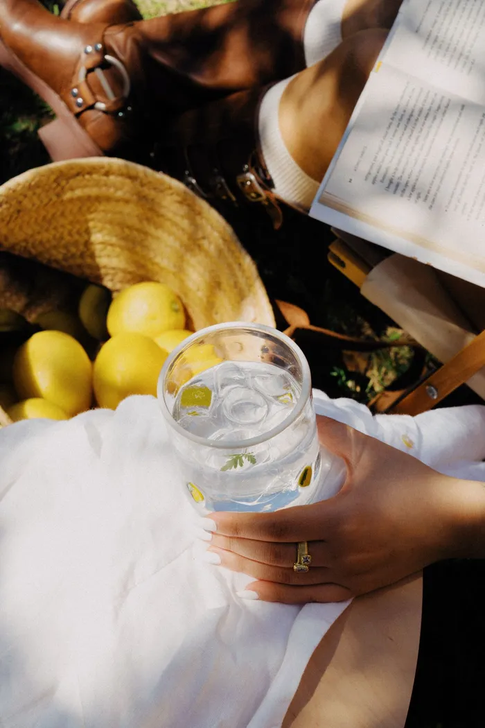 Hand holding a Lemon Icon Cup of iced water over white linen beside a basket of lemons