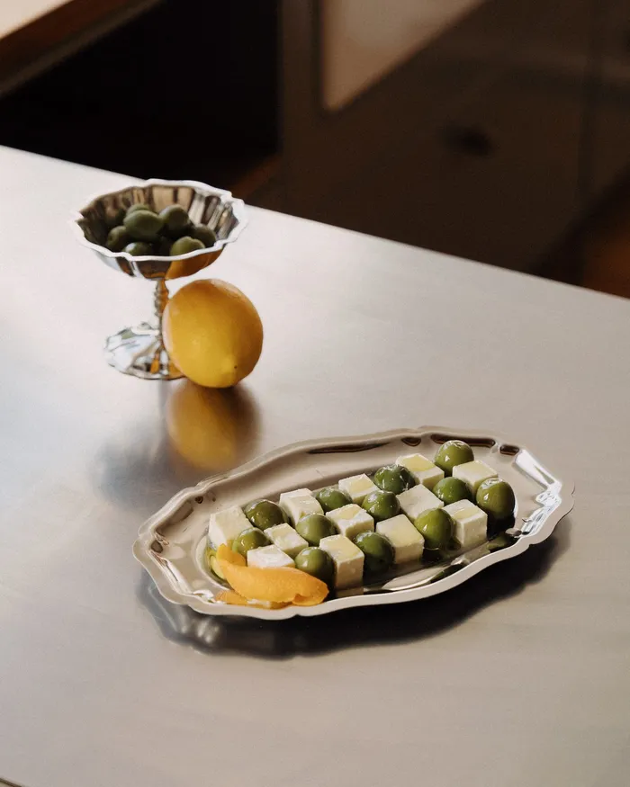 Riva coupe of olives beside a lemon and a Riva plate of olives and feta on a steel bench