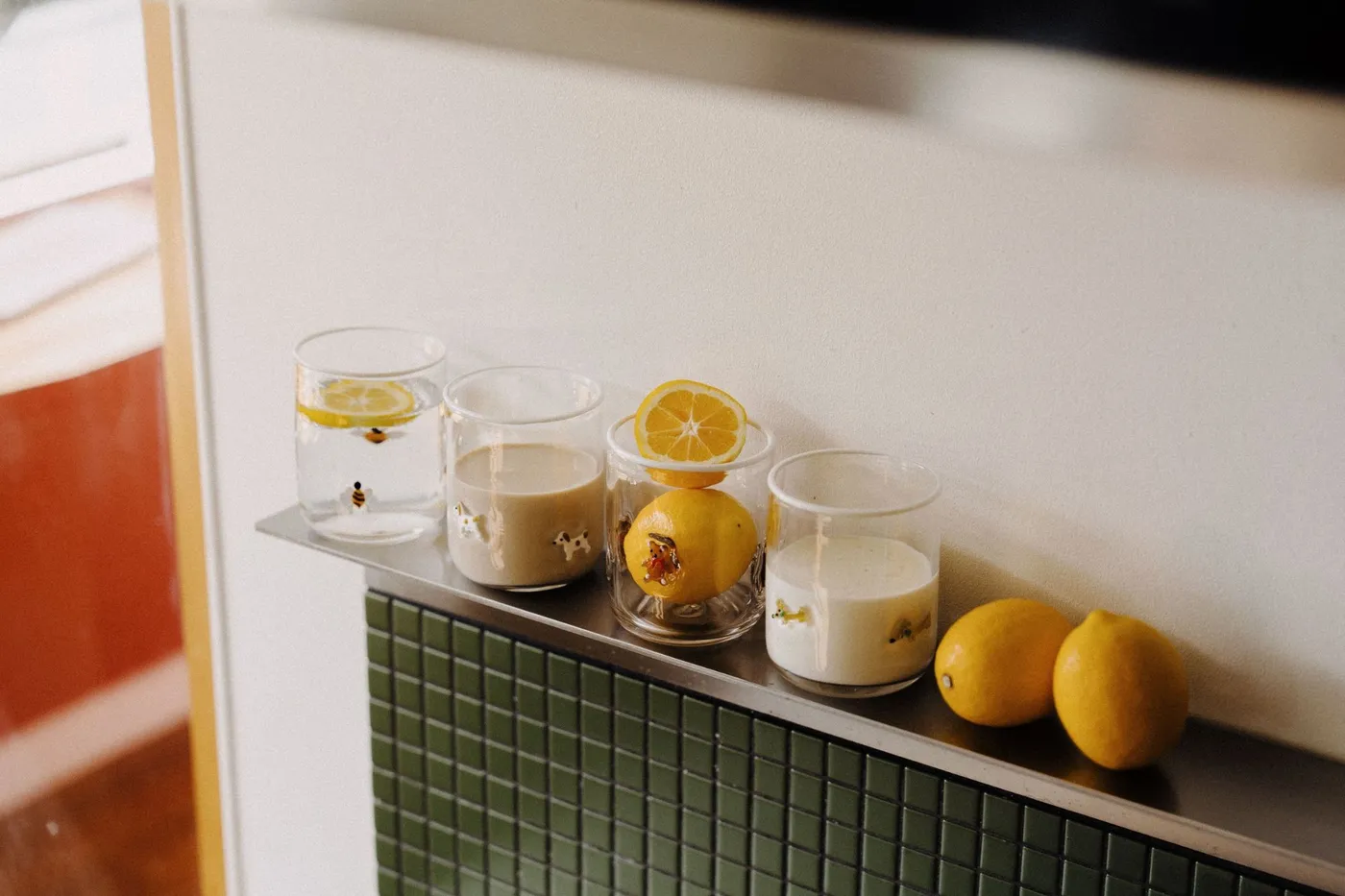 Four Icon Cups filled with water, milk and lemons on a steel shelf above green tiles